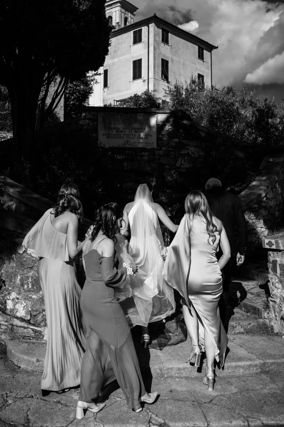 The bride and her bridesmaids climbing a stone staircase toward the villa, black and white photo