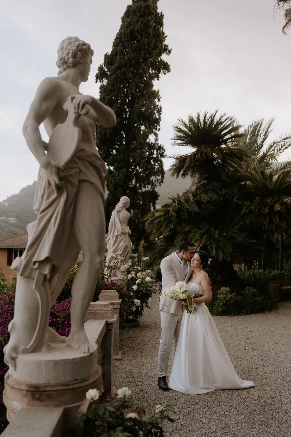 The couple embracing in the villa garden among statues and palm trees