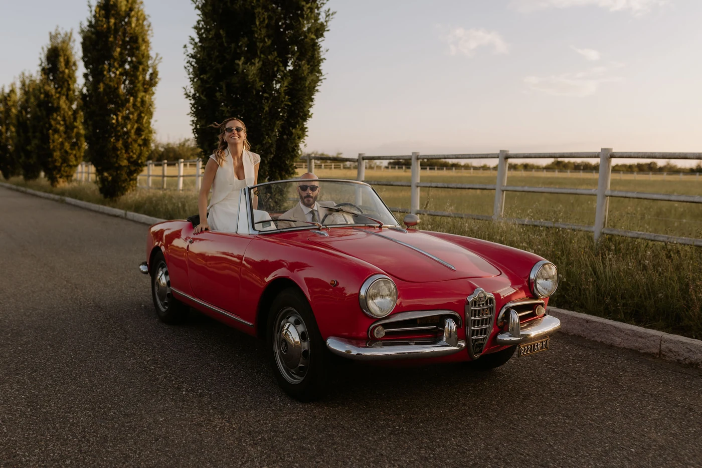 The bride standing in the red convertible driven by the groom, at sunset