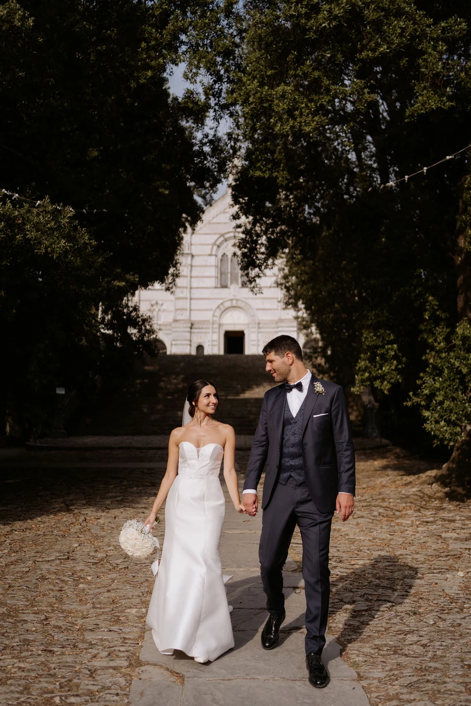 The couple walking hand in hand along the tree lined avenue in front of the church