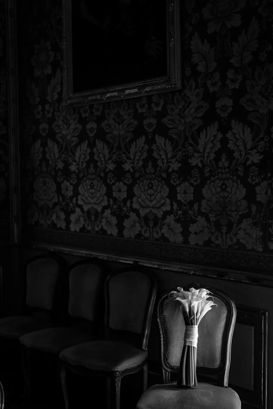 Bouquet of white calla lilies resting on an antique chair in a room with damask wallpaper, black and white photo