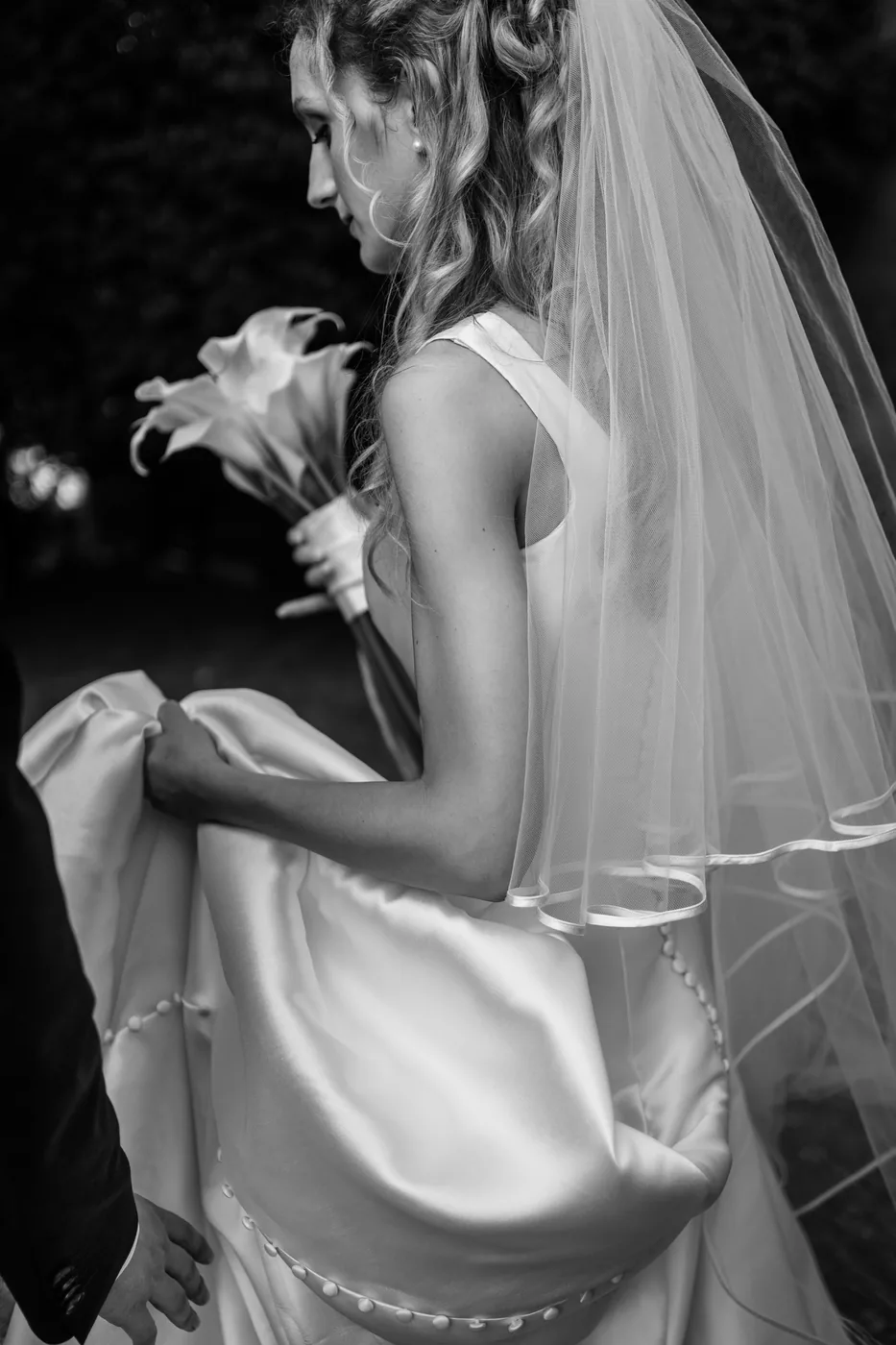 Bride in profile lifting her dress, holding a bouquet of calla lilies, black and white photo
