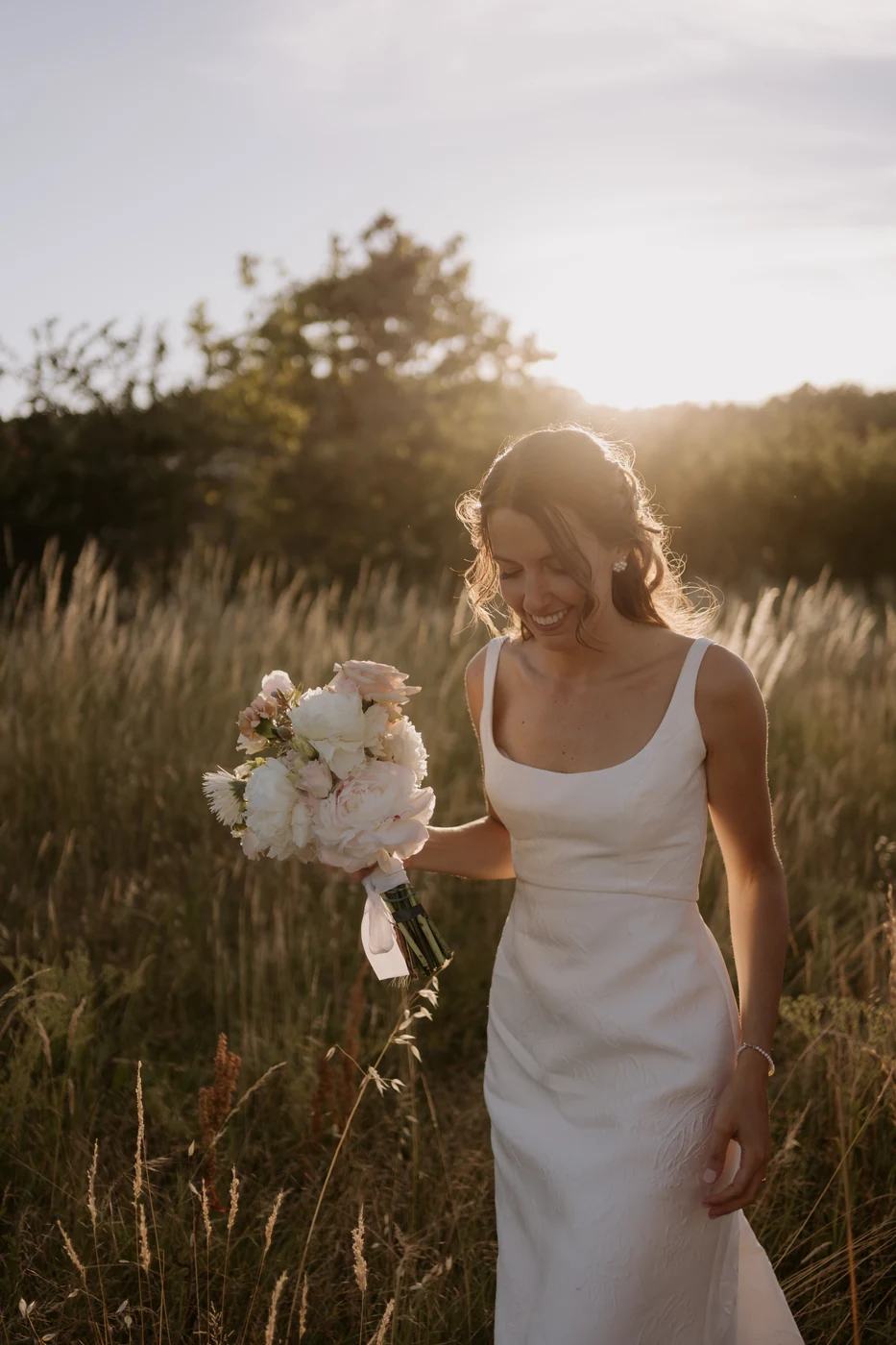 Smiling bride with her bouquet in the tall grass at sunset, wedding in Monferrato