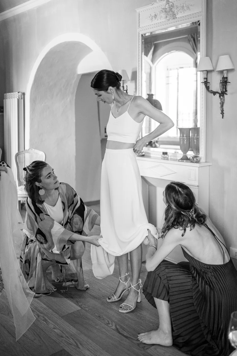 Friends helping the bride get dressed in front of the mirror, in black and white