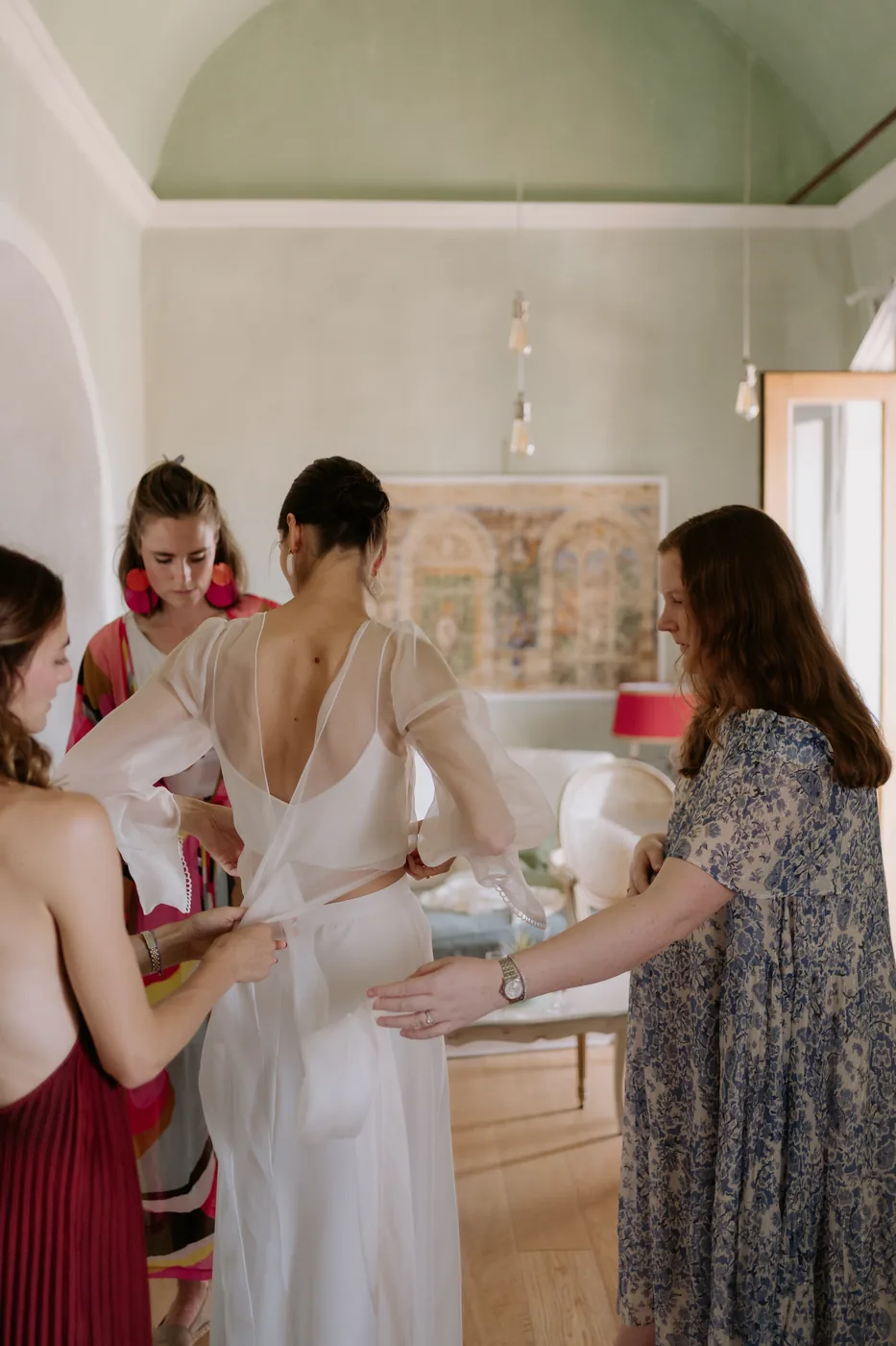 Friends fastening the bride's dress under the vaulted ceiling