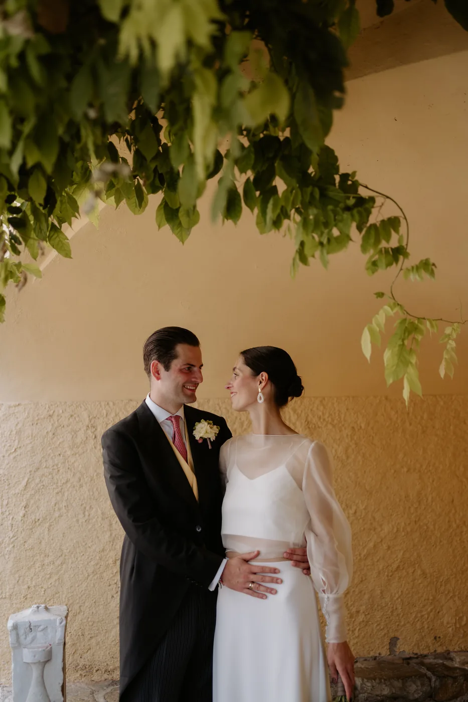 The couple embracing under the wisteria in front of a yellow wall