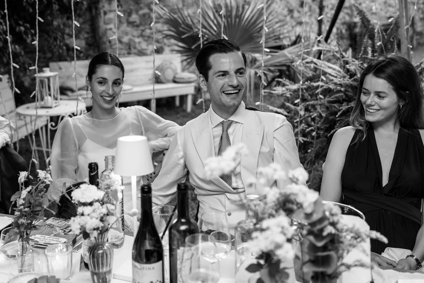 The groom laughing at the table between two guests, in black and white