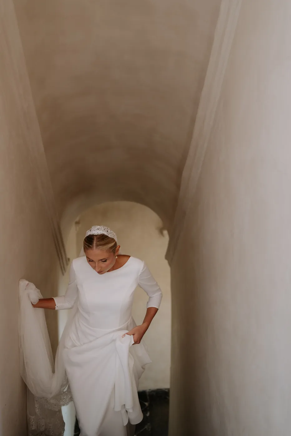 The bride walks down the stairs holding up her dress, veil on her head