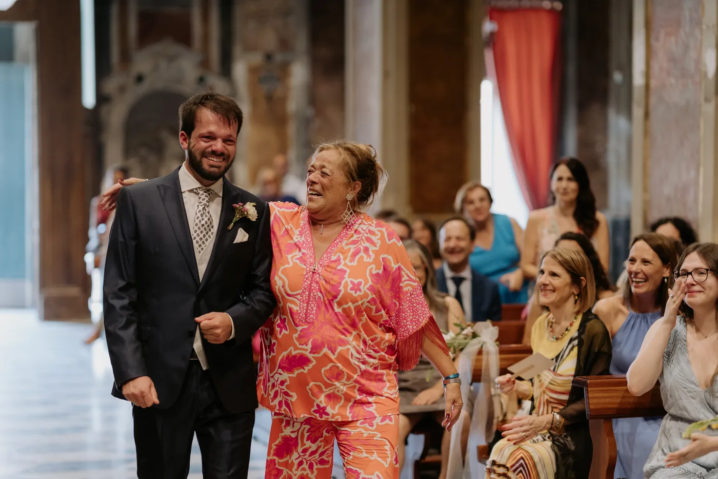 The groom entering the church arm in arm with a guest in a pink dress