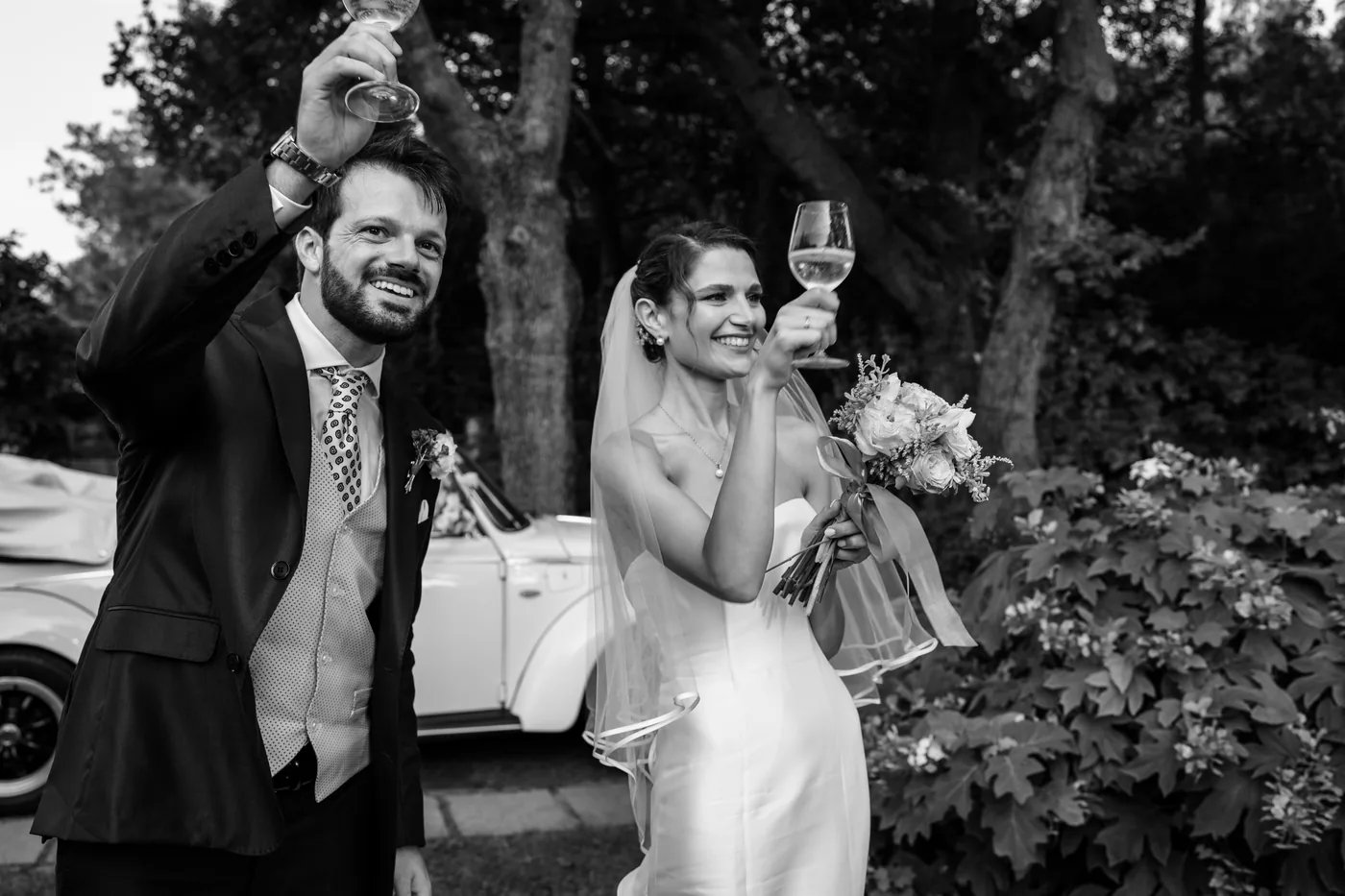 The groom waving as the bride laughs with a glass in her hand, in black and white