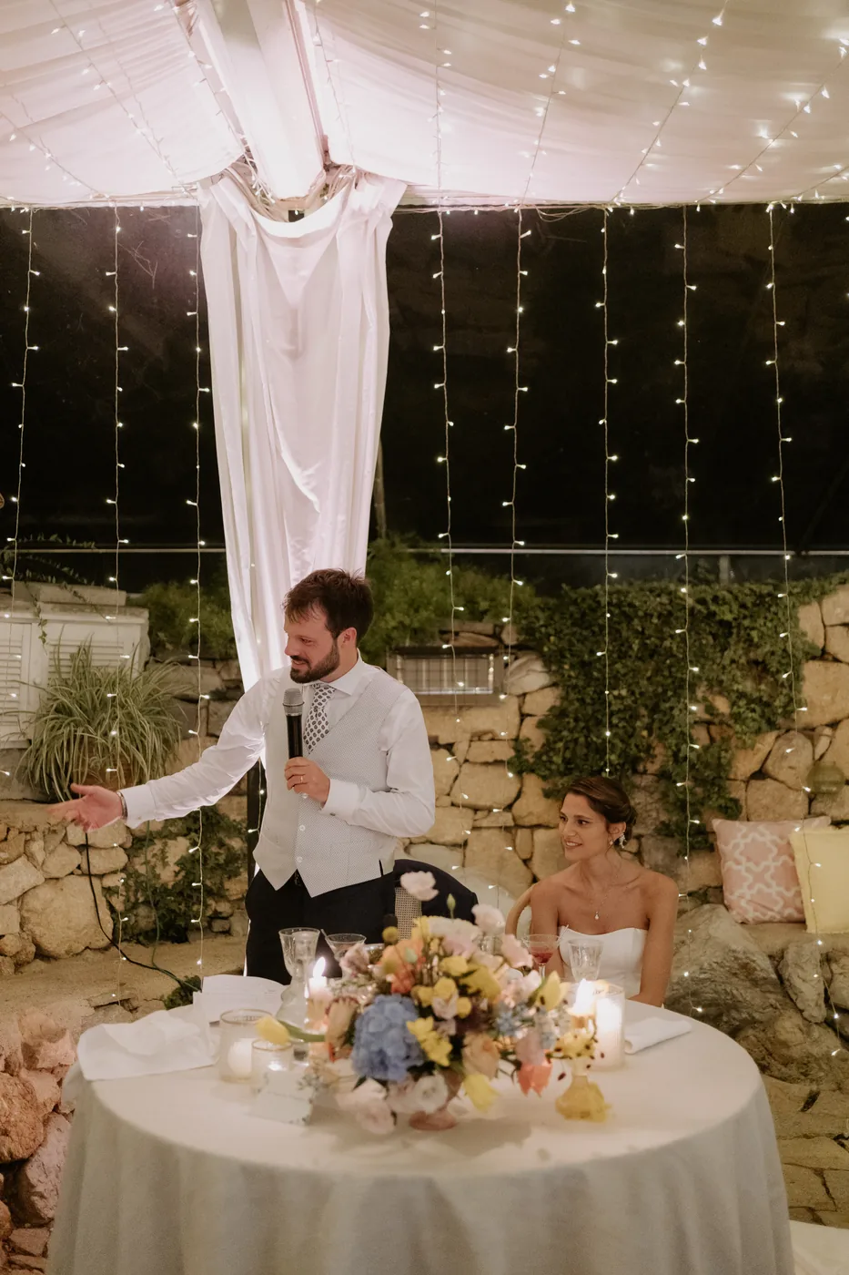 The groom giving a speech into the microphone beside the bride