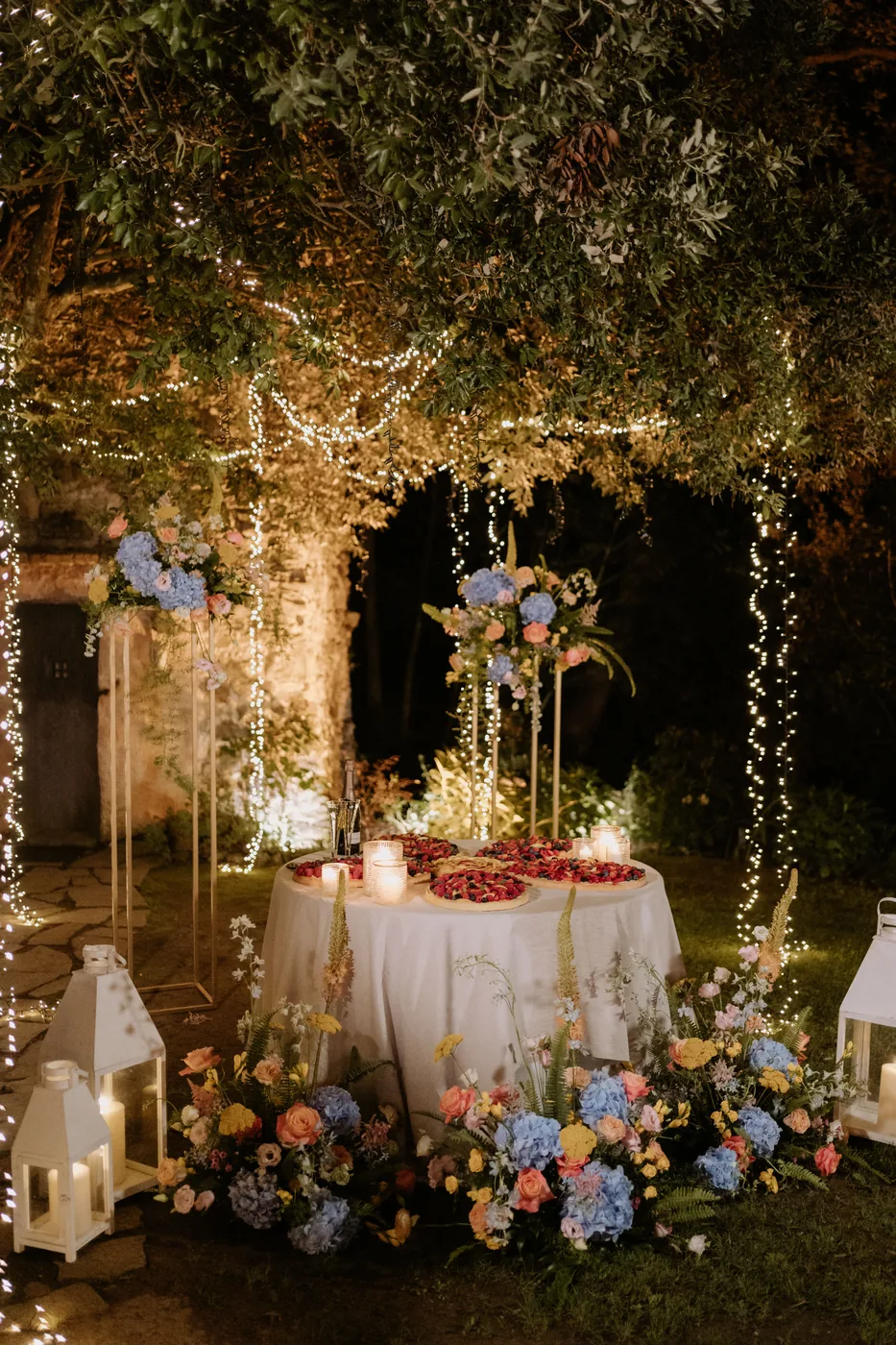 The cake table among flowers, lanterns and string lights in the garden